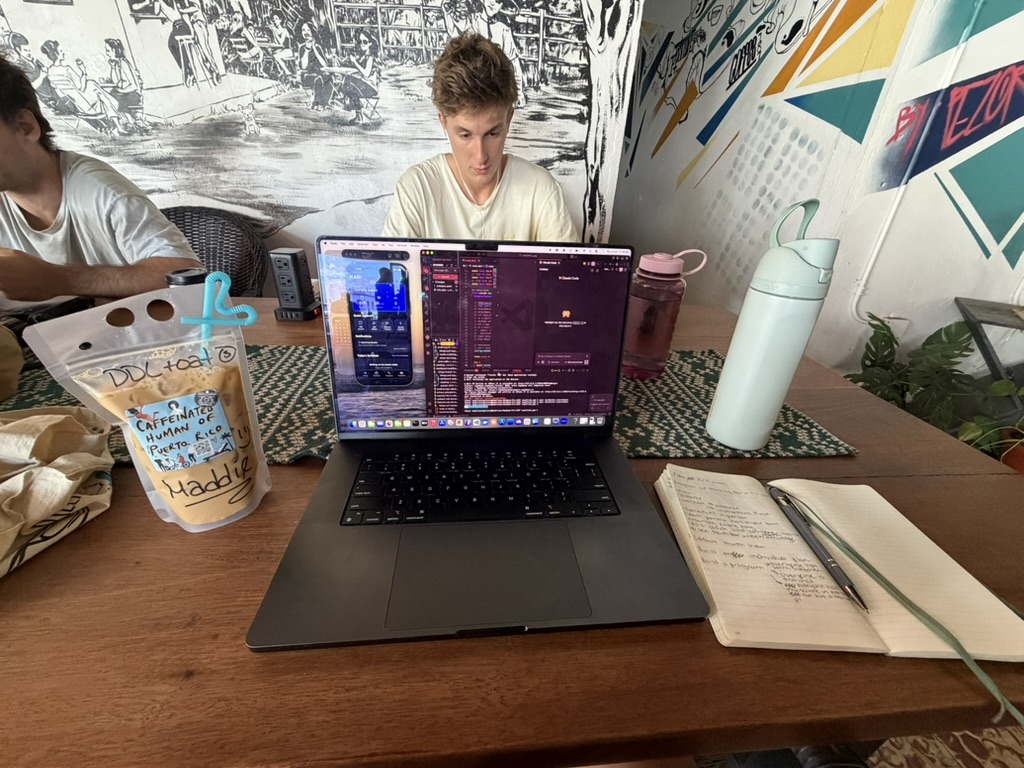 A laptop showing the KADI app and code on a café table beside drinks and a handwritten notebook.