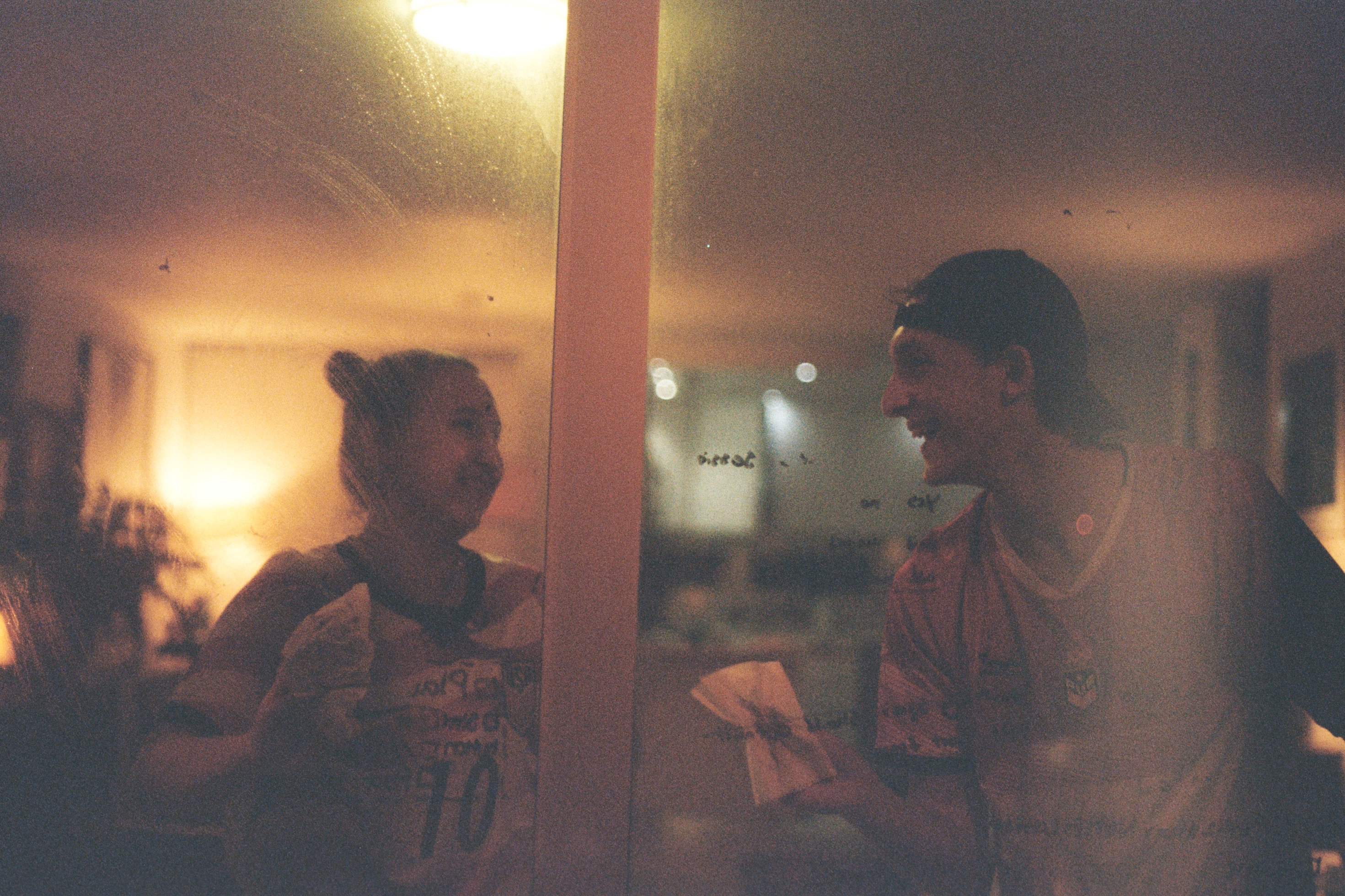 Maddie and Kit talking beside a warmly lit window.