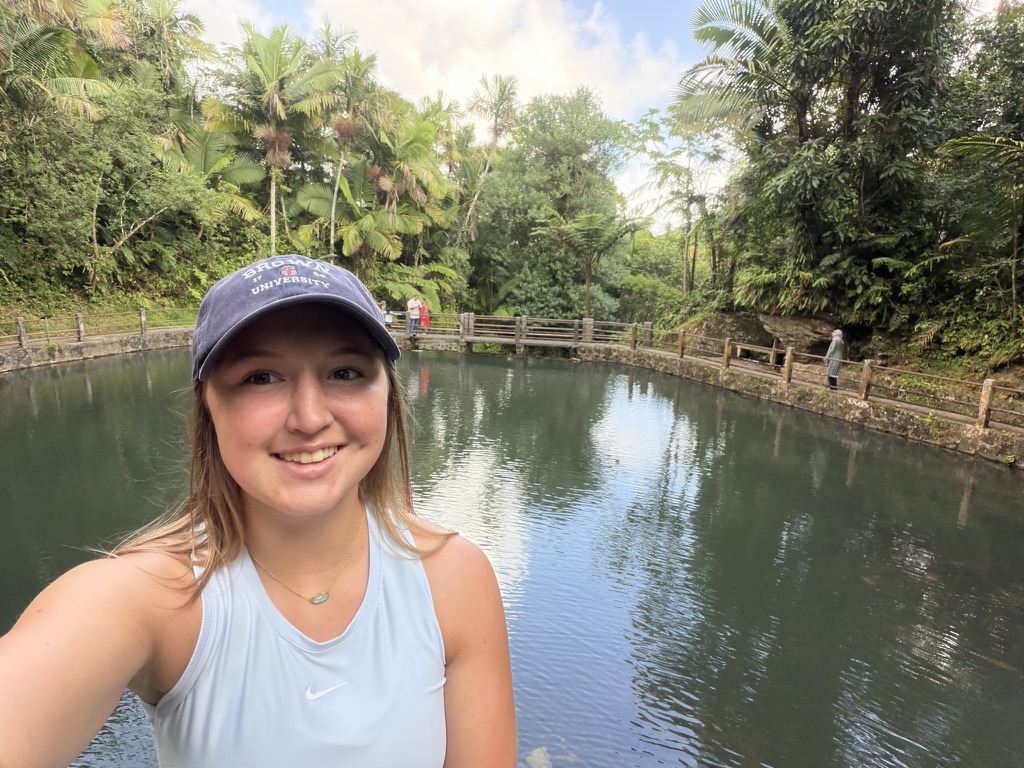 A selfie in a Brown University cap beside a pool surrounded by lush greenery.